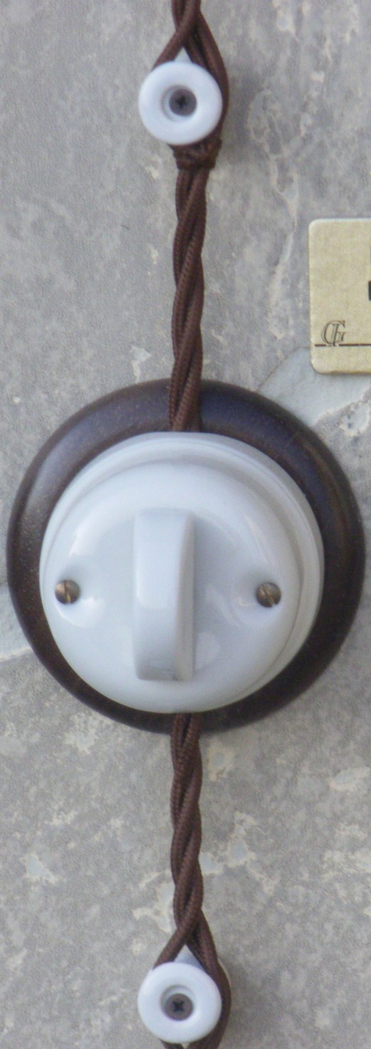 Porcelanowy włącznik światła natynkowy z brązową oprawką i kablem w brązowym oplocie, przymocowany do ściany za pomocą białych uchwytów.